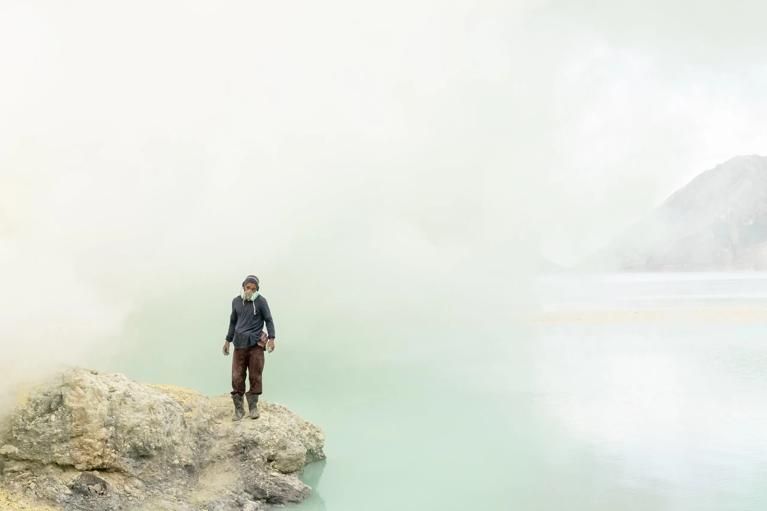 Lone miner standing on a rock at the milky turquoise acid lake of Kawah Ijen, shrouded in sulfur mist